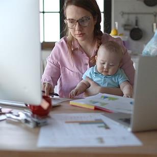 A mother is working at home, on her computer, while holding her baby.